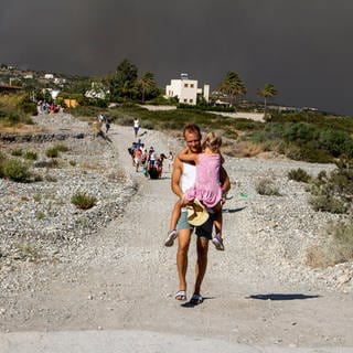 Ein Mann trägt ein Kind, als sie ein Waldbrandgebiet verlassen. Nach der Evakuierung vieler Dörfer und Hotelanlagen wegen großer Waldbrände haben Tausende Menschen auf der griechischen Ferieninsel Rhodos die Nacht im Freien verbracht. 