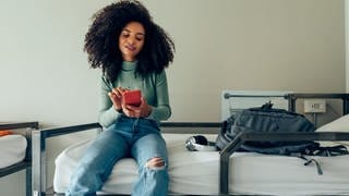 Das Handy als Schlüssel für Hiotelzimmer. Junge Frau sitzt mit einem Smartphone auf einem Hotelbett. 