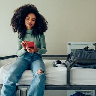 Das Handy als Schlüssel für Hiotelzimmer. Junge Frau sitzt mit einem Smartphone auf einem Hotelbett. 