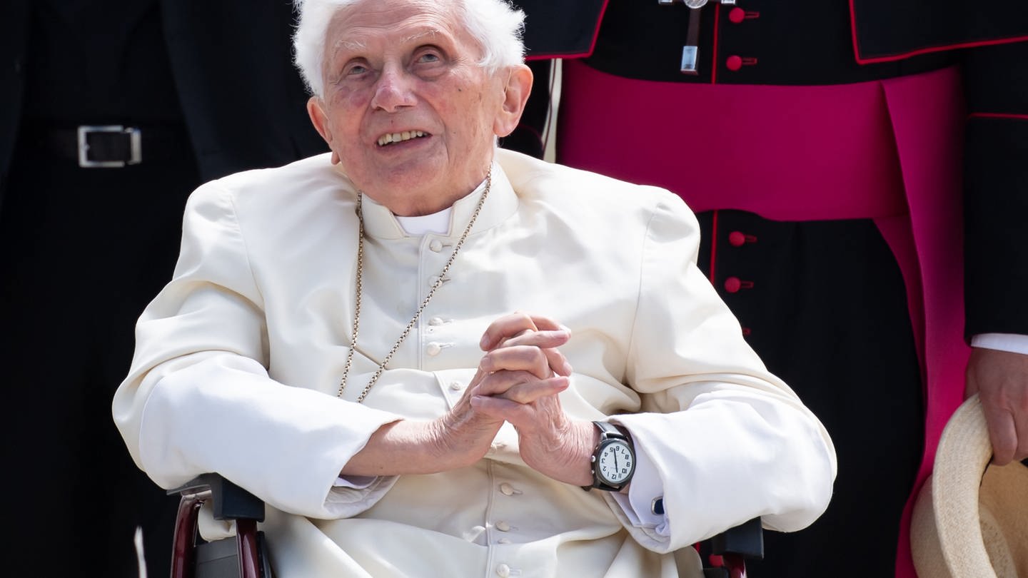 Der emeritierte Papst Benedikt XVI. sitzt mit gefalteten Händen in einem Rollstuhl.