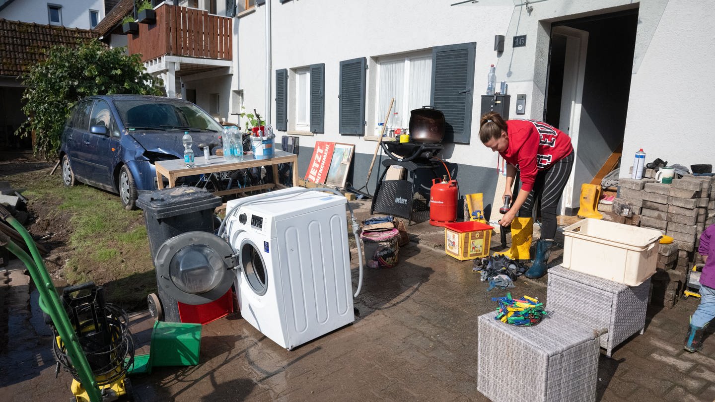 Aufräumen nach dem Hochwasser: Vor einem Haus stehen eine Waschmaschine, Möbel und ein Grill, eine Frau in Gummistiefeln wäscht Arbeitshandschuhe. Im Hintergrund steht ein schlammverschmutztes, beschädigtes Auto im Garten.