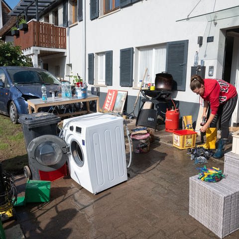 Aufräumen nach dem Hochwasser: Vor einem Haus stehen eine Waschmaschine, Möbel und ein Grill, eine Frau in Gummistiefeln wäscht Arbeitshandschuhe. Im Hintergrund steht ein schlammverschmutztes, beschädigtes Auto im Garten.