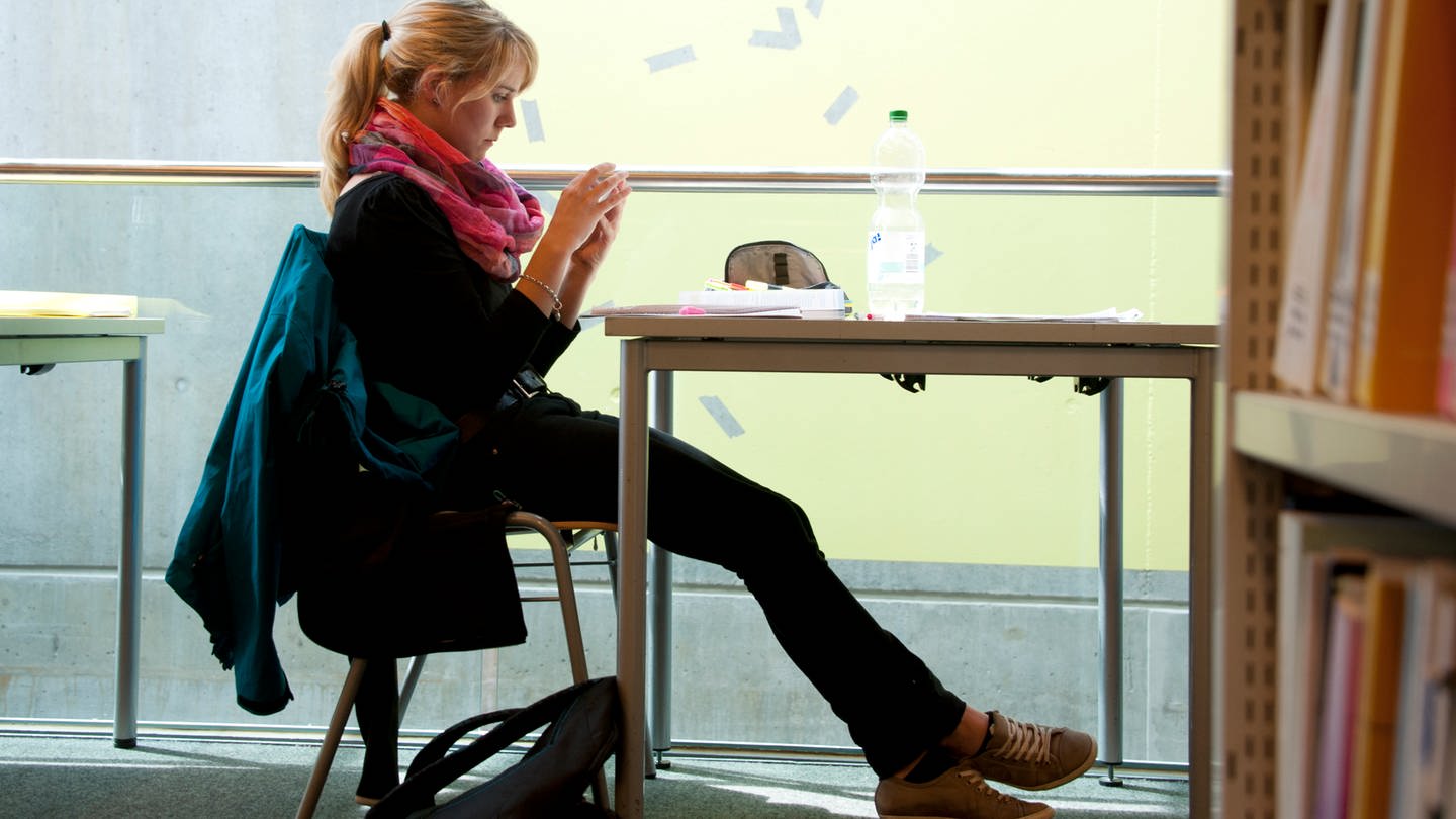 Eine Studentin schaut am 30.06.2014 in Tübingen (Baden-Württemberg) in der Uni-Bibliothek auf ihr Mobiltelefon. Langes Aufschieben von unliebsamen Aufgaben kann für Studenten zum Stress führen.