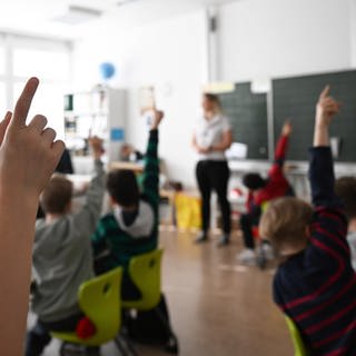 Viele Hände gehen in einem Klassenzimmer beim Unterricht an einer Grundschule in die Höhe. In der Ferne sieht man unscharf die Lehrerin.