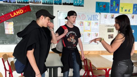Lehrerin steht rechts im Bild mit zwei Schülern links von ihr im Klassenzimmer und spricht mit ihnen.