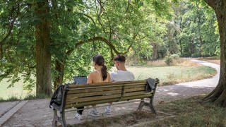 Zwei Personen sitzen im Schatten auf einer Bank. Die Person links arbeitet am Laptop.