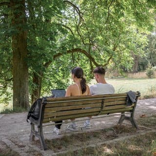 Zwei Personen sitzen im Schatten auf einer Bank. Die Person links arbeitet am Laptop.