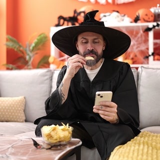 young bald man wearing wizard costume eating chips potatoes using smartphone at home