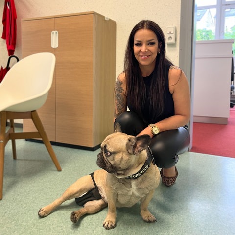 Lehrerin sitzt in der Hocke auf den Fußboden eines Klassenzimmers, vor ihr liegt ein Hund.