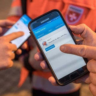 Zwei Rettungssanitäter bedienen die neue Ersthelfer-App Mobile Retter auf ihren Smartphones. Damit können sie nun auch außerhalb ihrer Dienstzeit per Smartphone-App alarmiert werden, um bei einem Herz-Kreislauf-Stillstand vor dem Eintreffen des Rettungsdienstes fachliche Hilfe leisten zu können.