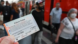 Symbolbild: Ein 9-Euro-Ticket für Juli 2022 wird am Hauptbahnhof hochgehalten, während Zugreisende aus einer Regionalbahn aussteigen.