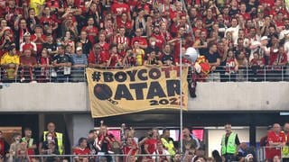 Die Freiburger Fankurve beim Spiel in der 1. Bundesliga gegen Borussia Dortmund am 12. August 2022. Über ein Geländer haben die Fans in roten Trikots ein Banner gehängt. Auf braunem Hintergrund steht in schwarzen Buchstaben "#Boycott Qatar 2022". Das Q ist geformt wie eine schwere Kugel an einer Kette. Die Fans protestieren gegen die hochumstrittene WM im Wüstenstaat Katar.  