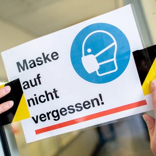 Eine Person klebt ein Schild mit der Aufschrift "Maske auf nicht vergessen!" an die Glasscheibe einer Zwischentür.