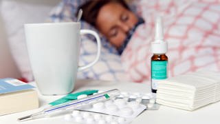 Eine Frau liegt mit Grippe im Bett, auf dem Nachtisch Fieberthermometer, Teetasse, Taschentücher, Tabletten und Nasenspray