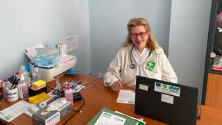 Irina von Zuboff sitzt im Arztkittel und mit Stethoskop um den Hals an einem Schreibtisch,