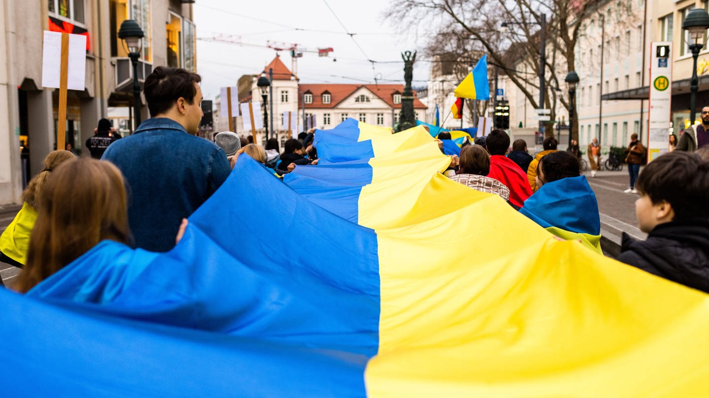 Menschen geehen während eines Marsches gegen den Angriffskrieg Russlands gegen die Ukraine durch die Innenstadt von Freiburg und halten dabei ein Banner in den ukrainischen Nationalfarben.
