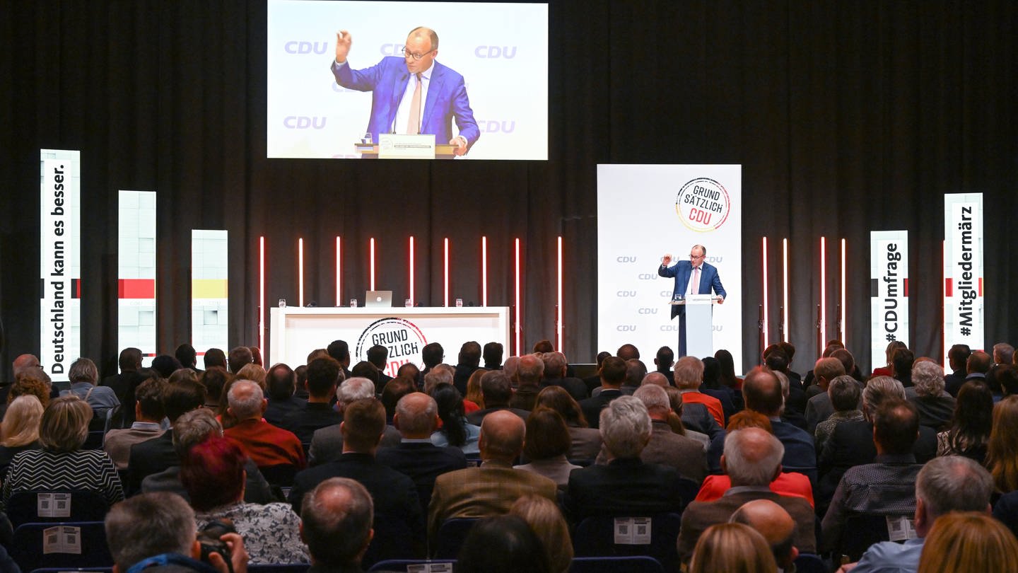 Ein Redner am Pult auf der Bühne, er wird auf eine Leinwand übertragen, im Vordergrund Publikum