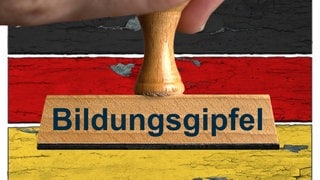 Ein symbolischer Holzstempel mit der Aufschrift "Bildungsgipfel", gehalten von einer Hand im Anschnitt vor einer Deutschland-Flagge auf drei rustikalen Brettern mit partiell abgeplatzer Farbe im Hintergrund.