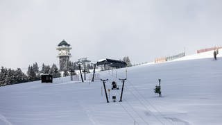 Schnee liegt auf einer Skispiste unter den Masten eines stillstehenden Lifts, während im Hintergrund der Feldbergturm zu sehen ist.
