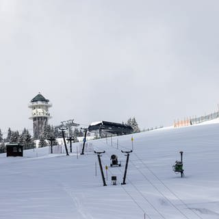 Schnee liegt auf einer Skispiste unter den Masten eines stillstehenden Lifts, während im Hintergrund der Feldbergturm zu sehen ist.