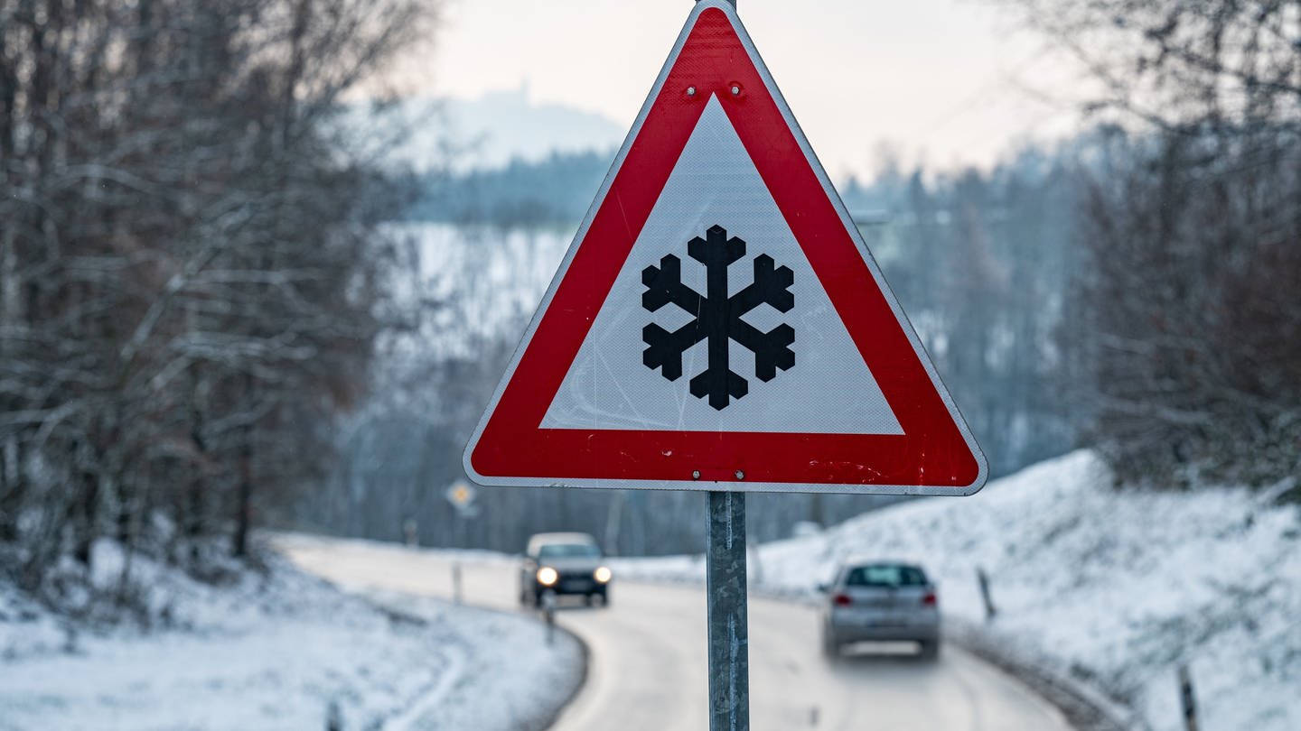 Ein Verkehrsschild mit einer Schneeflocke, das vor glatter Fahrbahn warnt steht am Straßenrand.