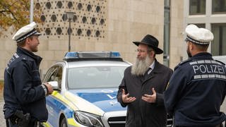 Rabbiner Shneur Trebnik steht mit zwei Polizisten vor der Synagoge an einem Polizeiauto.