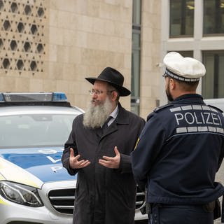 Rabbiner Shneur Trebnik steht mit zwei Polizisten vor der Synagoge an einem Polizeiauto.