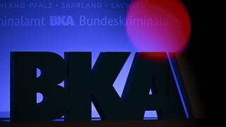 Das Rotlicht einer TV-Kamera leuchtet vor einem großen BKA-Logo.