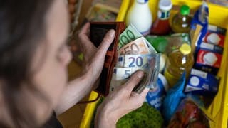 Eine Frau hält Geldscheine in einer Geldbörse über einer Einkaufskiste mit Lebensmitteln, der auf einem Küchentisch steht.