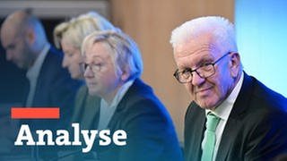 Ministerpräsident Winfried Kretschmann im rechten Bildrand schaut in die Kamera, im Hintergrund sein Kabinett.