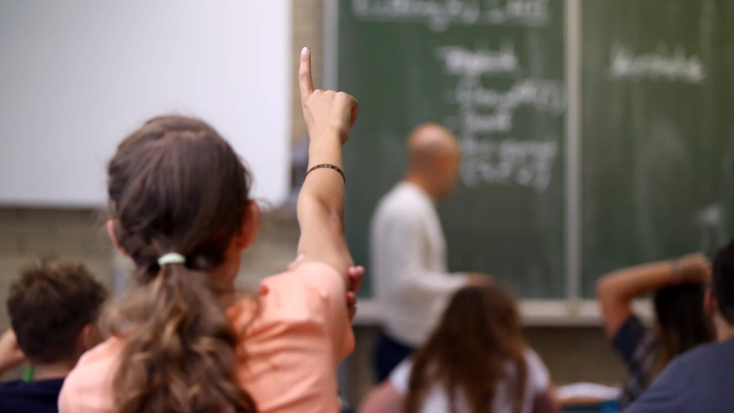 Eine Schülerin hebt die Hand, um sich zu melden. Sie sitzt im Klassenzimmer vor einer Tafel.