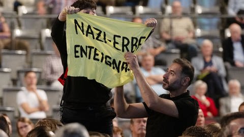 Eine Frau hält während der Porsche Hauptversammlung ein Transparent mit der Aufschrift „Nazi-Erbe enteignen“.