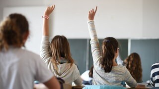 Zwei Schülerinnen strecken in einem Klassenzimmer ihre Hände nach oben. 