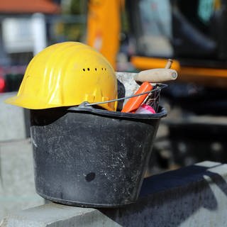 In Baden-Württemberg fehlen rund 70.000 Wohnungen - schätzt der Lobbyverband der Bauwirtschaft.Ein gelber Bauarbeiterhelm liegt auf einem Eimer eines Maurers auf einer Baustelle.