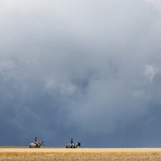 Reiterinnen sind mit ihren Pferden zwischen Getreidefeldern unterwegs, während im Hintergrund ein Gewitter aufzieht.