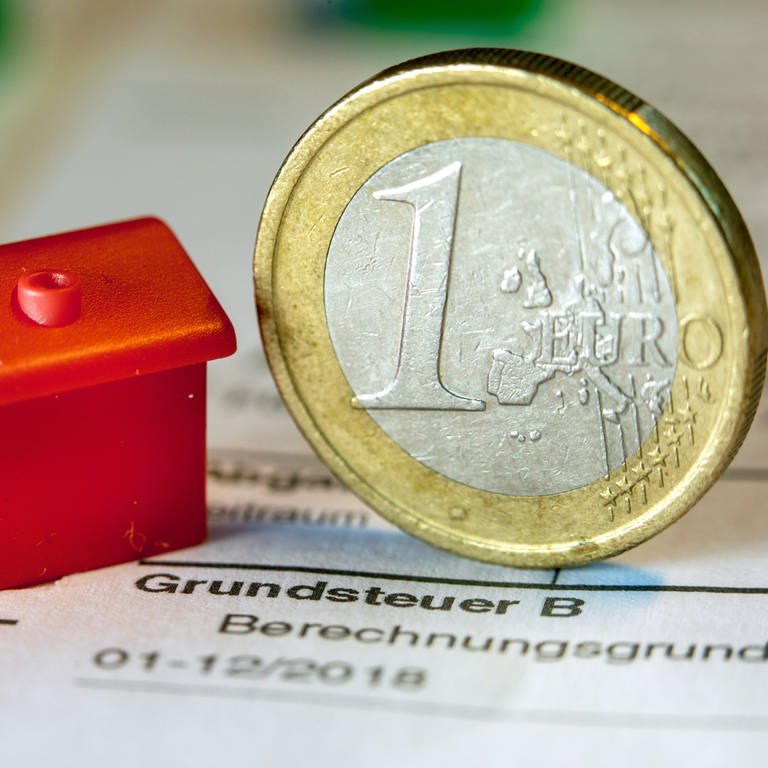 Eine Euro-Geldmünze sowie ein Spielzeughaus stehen auf einem Abgabenbescheid für die Entrichtung der Grundsteuer.