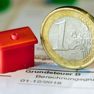 Eine Euro-Geldmünze sowie ein Spielzeughaus stehen auf einem Abgabenbescheid für die Entrichtung der Grundsteuer.