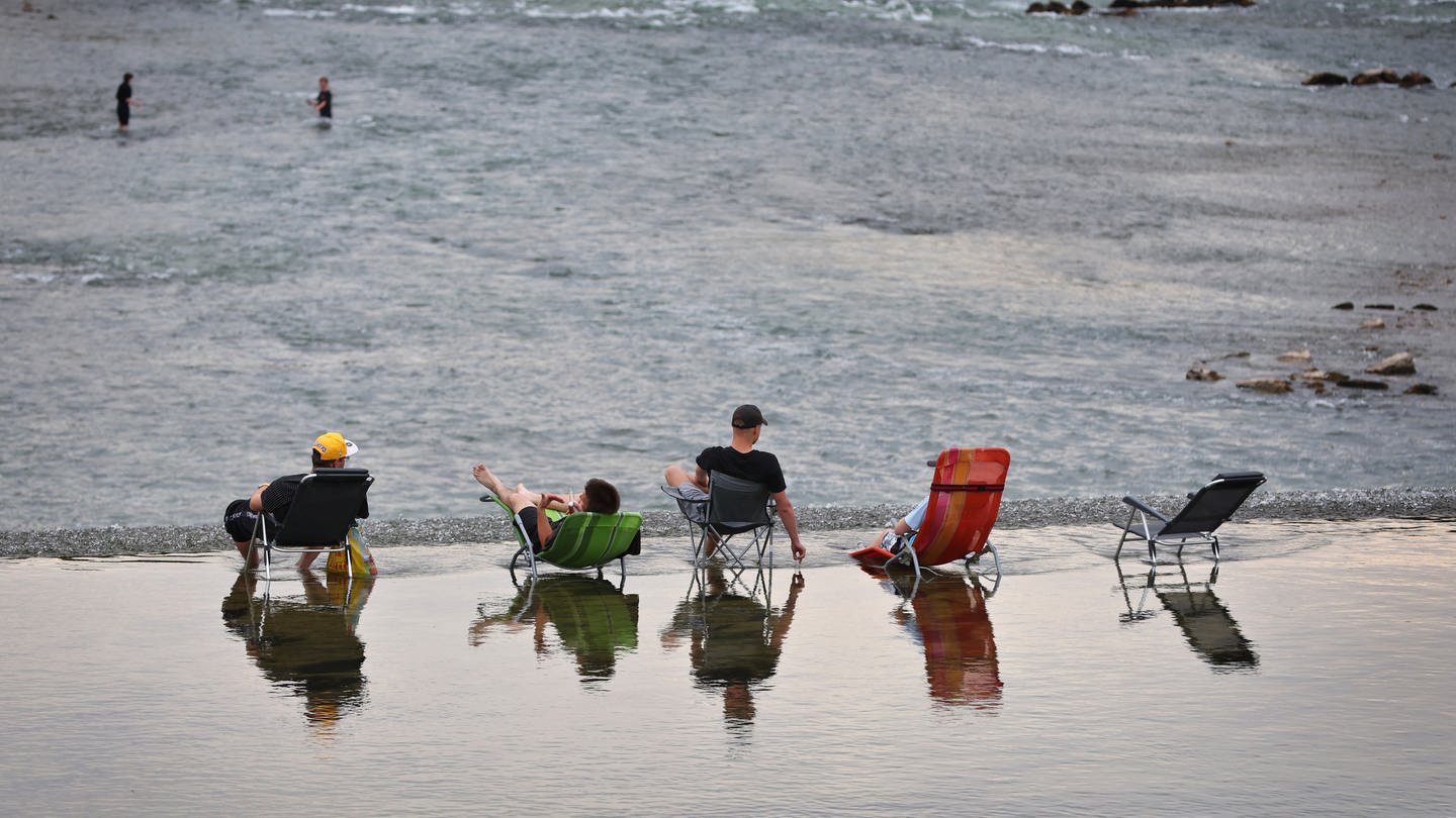 Junge Männer sitzen auf Campingstühlen im seichten Wasser oberhalb des Lechwehrs.