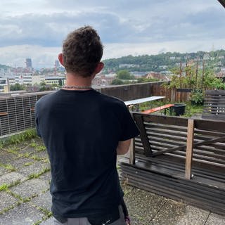 Ein junger Mann in Arbeitshose und schwarzem T-Shirt von hinten fotografiert schaut über die Dächer von Stuttgart-Feuerbach. 