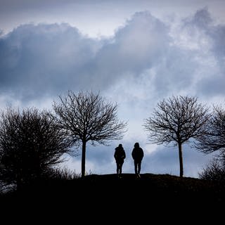 Spaziergänger gehen auf einen Hügel am Kronsberg, während bei stürmischem Wetter dunkle Wolken am Horizont vorüberziehen.