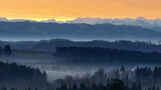 Die Sonne geht hinter dem Horizont über den Alpen auf (Aufnahme mit Drohne). Leichter Bodennebel bedeckt die Landschaft im Voralpenland.