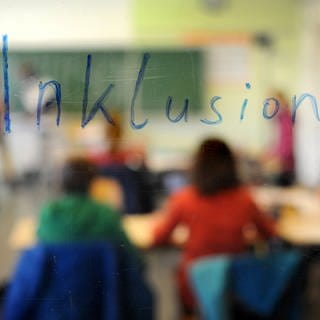 Hinter einer Glasscheibe mit der Aufschrift "Inklusion" sitzen Schülerinnen und Schüler in einem Klassenzimmer. 