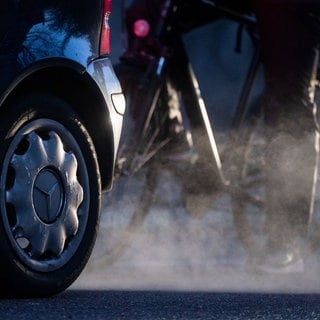 Ein Radfahrer steht neben einem Auto von Mercedes mit Dieselantrieb, dessen Abgase in der kalten Morgenluft sichtbar werden.