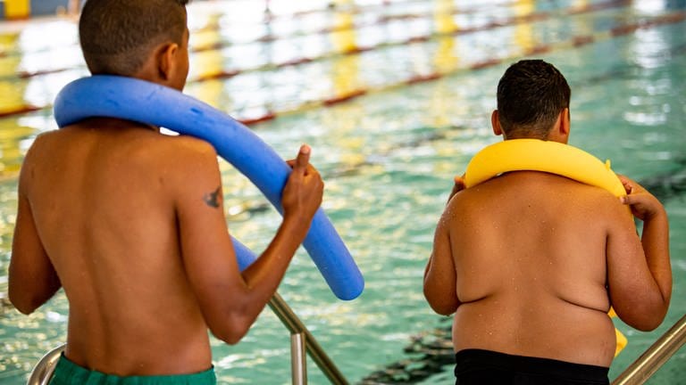 Zwei Kinder stehen in einem Hallenbad mit einer Schwimmnudel am Beckenrand - eines davon ist übergewichtig. (Symbolbild)