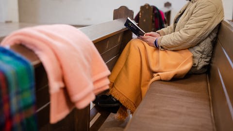 Eine Frau sitzt mit einer Decke in der Kirche. Auch Kirchen müssen wegen der Energiekrise unter anderem beim Heizen sparen.