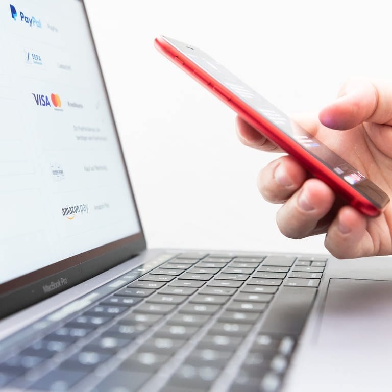 Ein Mann hält sein Smartphone zur Zwei-Faktoren-Authentifizierung in der Hand, während auf dem Bildschirm eines Laptops verschiedene Bezahlmethoden abgebildet sind. 