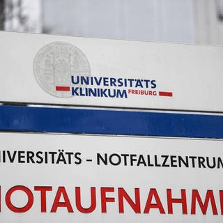 Ein Schild mit der Aufschrift "Universitätsklinikum Freiburg und Notaufnahme" steht vor einem Gebäude.