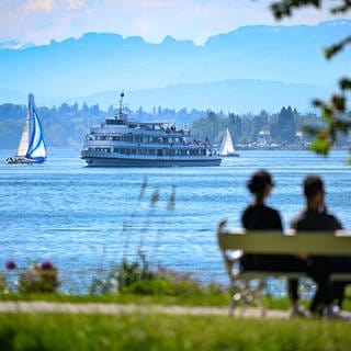Das Passagierschiff Stuttgart fährt auf dem Bodensee zur Anlegestelle, während im Vordergund ein Paar auf einer Bank im Uferpark sitzt.