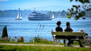 Das Passagierschiff Stuttgart fährt auf dem Bodensee zur Anlegestelle, während im Vordergund ein Paar auf einer Bank im Uferpark sitzt.