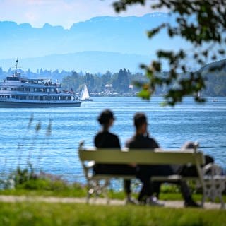 Das Passagierschiff Stuttgart fährt auf dem Bodensee zur Anlegestelle, während im Vordergund ein Paar auf einer Bank im Uferpark sitzt.